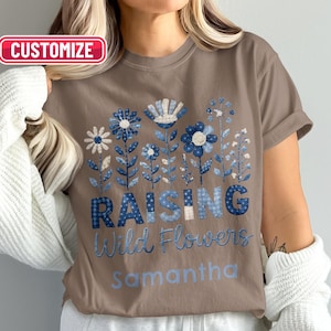 Puede incluir: Una camiseta marrón de manga corta con un gráfico de flores azules y blancas estampadas. Debajo de las flores, el texto dice "RAISING Wild Flowers" en azul, seguido del nombre "samantha" en azul claro. Se ve una etiqueta "CUSTOMIZE".