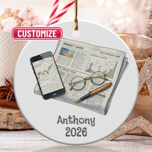 May include: A round, white ceramic ornament featuring a newspaper, glasses, a pen, and a smartphone displaying a stock chart. The ornament has the text "Anthony 2026" and a red "Customize" label.