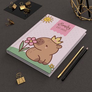 May include: A personalised journal featuring a cartoon capybara wearing a crown and smelling a flower. The cover is pink with a sun and the name "Emily" and "EST 2010". Gold and black pencils and gold binder clips are nearby.