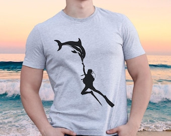 Może przedstawiać: Jasnoszary t-shirt z krótkim rękawem z czarnym nadrukiem graficznym przedstawiającym nurka sięgającego do płetwy skaczącego delfina. Wzór jest wyśrodkowany na klatce piersiowej odzieży, która posiada standardowy okrągły dekolt.