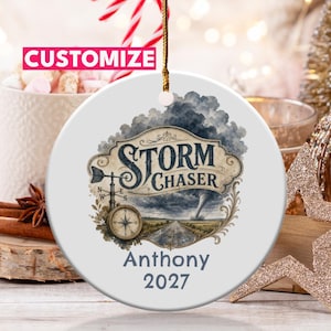 Op de afbeelding: Een rond wit keramieken ornament met een afbeelding van een storm met een tornado, een kompas en een windwijzer. De tekst luidt STORM CHASER, Anthony en 2027. Het ornament hangt aan een gouden koord en bevat een roze CUSTOMIZE-label.