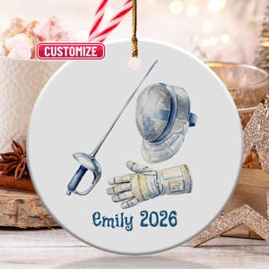 May include: A white ceramic ornament featuring a watercolor illustration of fencing equipment: a foil, mask, and glove. The text "Emily 2026" is printed in blue script. The ornament is hanging with a gold string, with a "Customize" button.