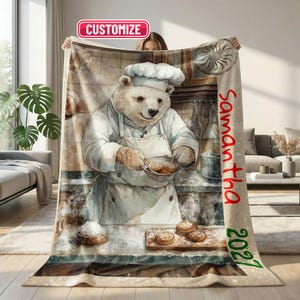 Puede incluir: Una manta suave con un oso blanco con gorro y delantal de chef, sosteniendo un cuenco de galletas en una cocina. El texto CUSTOMIZE aparece arriba, mientras que el nombre Samantha y el año 2027 están impresos en rojo en el lado derecho.