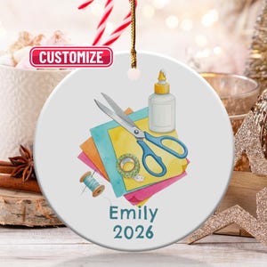 May include: A white ceramic ornament featuring a watercolor illustration of craft supplies. The design includes blue scissors, glue, colorful paper, washi tape, and a spool of thread. The name "Emily" and the year "2026" are printed in teal. A gold string is attached for hanging.