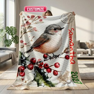 May include: A soft throw blanket featuring a watercolour illustration of a small bird perched on a holly branch with red berries. The text "CUSTOMIZE" appears at the top, while the name "Samantha" and the year "2027" are printed in red on the side.