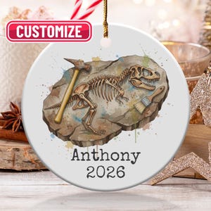 May include: A white ceramic ornament featuring a watercolor illustration of a dinosaur skeleton fossil with a hammer and brush. The ornament is personalized with the name "Anthony" and the year "2026". The word "Customize" is in a red box at the top.