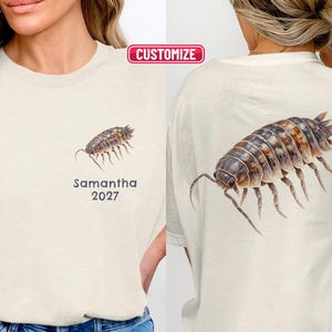 Può includere: Una t-shirt color crema con una piccola illustrazione di un porcellino di terra sul petto sinistro con il testo Samantha 2027 sotto, e una grande grafica dettagliata di un porcellino di terra stampata sul retro del capo.