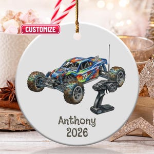 May include: A white ceramic ornament featuring a colorful illustration of a remote-controlled monster truck. The ornament includes the text "Anthony 2026" and a black remote control. The ornament is round and has a gold string for hanging.