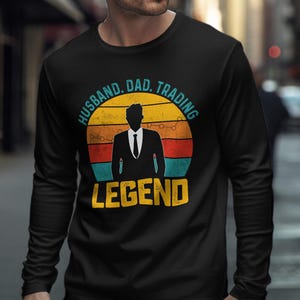 May include: A black long-sleeve shirt featuring a graphic of a silhouette in a suit against a sunset background. The text reads HUSBAND. DAD. TRADING LEGEND in a circular arrangement around the graphic, with the word LEGEND in large yellow letters.