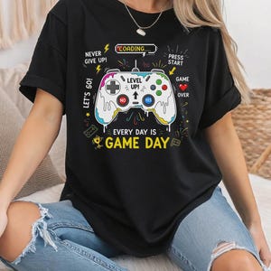 Puede incluir: Camiseta negra con un diseño de mando de videojuegos colorido. El diseño incluye los textos "NEVER GIVE UP!", "LET'S GO!", "LEVEL UP!" y "EVERY DAY IS GAME DAY". El mando es blanco con botones coloridos.