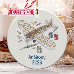 May include: A round, white ceramic ornament featuring a detailed illustration of a model airplane with tools and paint. The text "Anthony 2026" is printed in blue. A gold string is attached for hanging.