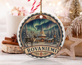 May include: A round white ceramic ornament featuring a winter scene with a reindeer, a snow-covered building, and the aurora borealis in the sky. The word ROVANIEMI is printed in dark blue capital letters on a banner at the bottom of the design.