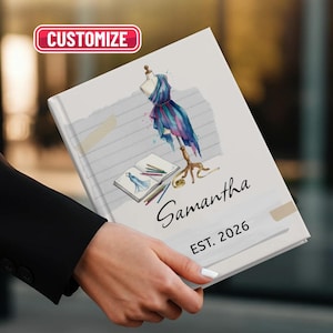 Può includere: Un taccuino personalizzato a tema fashion design. La copertina presenta un'illustrazione ad acquerello di un manichino con un abito blu e viola, un taccuino e delle matite. Il nome "Samantha" e "EST. 2026" sono stampati.