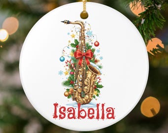 May include: A white, round Christmas ornament featuring a gold saxophone decorated with a red bow, Christmas lights, and ornaments. The name "Isabella" is written in red script at the bottom. A gold string is attached for hanging.