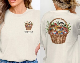 May include: Off-white t-shirt with a watercolour illustration of a wicker basket filled with mushrooms, berries, and foliage. The name "EMILY" is printed below the basket on the front. The back of the shirt also displays the basket design.