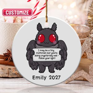 May include: A round white ceramic ornament featuring a cartoon black mothman with large red eyes holding a sign that reads, I may be a tiny mothman but you're doing cryptid-ibly well - follow your light! The name Emily and year 2027 are printed below.