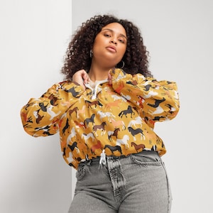May include: A mustard yellow quarter-zip pullover jacket featuring an all-over print of various horse breeds in shades of brown, black, white, and tan. The jacket has long sleeves, a white front zipper, and a white drawstring at the bottom hem.