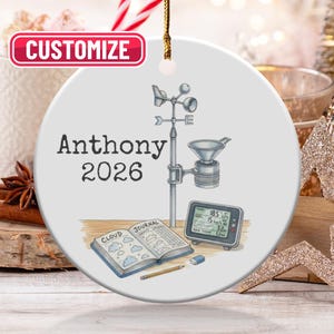 Puede incluir: Adorno navideño redondo de cerámica blanca con hilo dorado. Presenta una ilustración de estación meteorológica, un diario y el nombre "Anthony" con el año "2026". La palabra "Customize" está en un cuadro rojo en la parte superior.