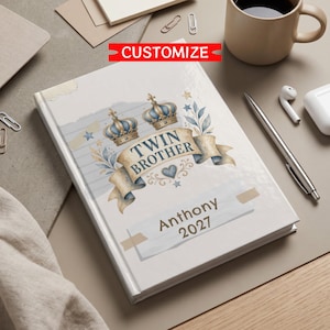 May include: A hardback journal featuring a cream-coloured cover with a graphic of two blue and gold crowns above a ribbon banner that reads TWIN BROTHER. Below the banner, the name Anthony and the year 2027 are printed in dark brown text.