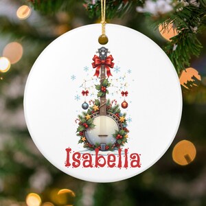 May include: A white ceramic Christmas ornament featuring a banjo decorated with festive holiday elements. The banjo is adorned with a red bow, holly, lights, and ornaments. The name "Isabella" is written in red script at the bottom.