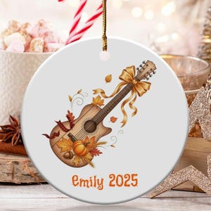 May include: A round, white ceramic ornament featuring a watercolor guitar design with autumn leaves, a pumpkin, and a gold bow. The text "Emily 2025" is printed in orange. A gold string is attached for hanging.