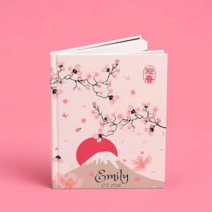 Puede incluir: Un libro de tapa dura con una cubierta de color rosa claro que presenta un gráfico de un sol rojo, una montaña y ramas de cerezo. El texto "Emily" y "EST 2004" está impreso en la parte frontal, junto a un sello circular con los caracteres 迎春.