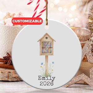 May include: A white ceramic Christmas ornament featuring a watercolor illustration of a miniature library. The ornament has a gold string for hanging and includes the name "Emily" and the year "2026". The word "CUSTOMIZABLE" is in a red oval.