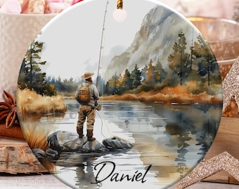 May include: A round ceramic Christmas ornament featuring a watercolor painting of a fisherman standing on a rock in a lake. The scene includes mountains, trees, and the name "Daniel" in script. The ornament has a gold string for hanging.