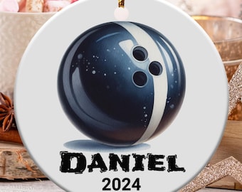 May include: A round, white ceramic ornament featuring a navy blue bowling ball design with a white stripe and three finger holes. The name "DANIEL" and the year "2024" are printed in black below the bowling ball.