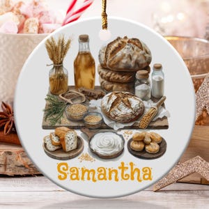 May include: A round ceramic ornament featuring a bread-themed illustration. The design includes various types of bread, grains, and ingredients, with the name "Samantha" in orange script at the bottom. A gold string is attached for hanging.