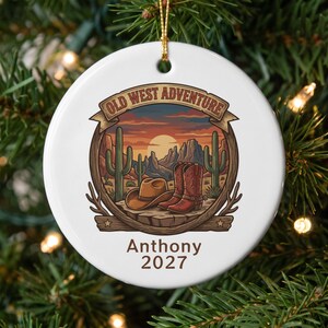 May include: A round white ceramic ornament featuring a desert landscape illustration with a cowboy hat, red boots, and cacti. The text reads OLD WEST ADVENTURE at the top and Anthony 2027 at the bottom. It hangs from a gold string.