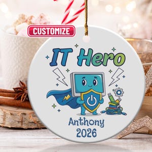 Puede incluir: Adorno navideño de cerámica blanca con un diseño de héroe informático de dibujos animados. El diseño presenta un personaje de pantalla de ordenador con capa y escudo. También se muestra el texto "IT Hero", el nombre "Anthony" y el año "2026".