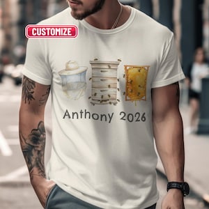 May include: White t-shirt with a bee-themed design. The design includes a beekeeper's hat, a beehive, and a honeycomb, with the name "Anthony" and the year "2026" printed below. The word "Customize" is in a red box at the top.