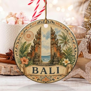 Peut inclure: Une décoration ronde en céramique illustrant une porte de temple balinais, des palmiers et des vagues. Le mot BALI est inscrit en lettres capitales noires sur une bannière décorative en bas, avec une ficelle dorée pour la suspension.