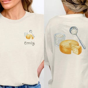 Op de afbeelding: Crèmekleurig T-shirt met een aquarel illustratie van kaas en een lepel. De voorkant van het shirt heeft de naam "Emily" in script. De achterkant van het shirt heeft een grotere kaas illustratie.