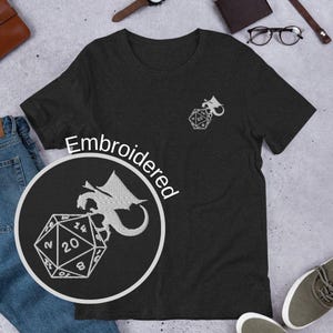 May include: Dark gray t-shirt with an embroidered design of a dragon and a twenty-sided die. The word "Embroidered" is printed on the shirt. A smaller version of the design is on the upper right chest.