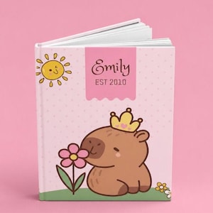 Könnte beinhalten: Ein Hardcover-Buch mit hellrosa Einband und einem Muster aus kleinen Punkten. Auf dem Einband ist ein Cartoon-Wasserschwein mit gelber Krone, eine rosa Blume, eine gelbe Sonne und ein rosa Banner mit dem Text Emily EST 2010 in Dunkelbraun.
