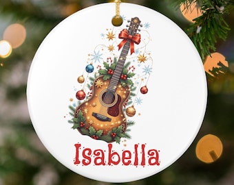May include: A white, round Christmas ornament featuring a detailed illustration of an acoustic guitar decorated with Christmas ornaments, a red bow, and holly. The name "Isabella" is written in red script at the bottom. A gold string is attached for hanging.