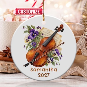 May include: A round white ceramic ornament featuring a brown violin with a bow, purple and yellow pansy flowers, and sheet music. The text "Samantha" and "2027" is printed in brown below the illustration. A gold string is attached at the top.