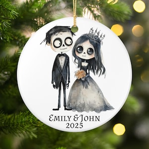May include: A white ceramic Christmas ornament featuring a gothic wedding couple illustration. The bride wears a black dress and crown, holding a bouquet. The groom is in a black tuxedo. The text "EMILY & JOHN 2025" is at the bottom.