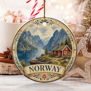 Puede incluir: Un adorno redondo de cerámica con una ilustración escénica de una cabaña roja junto a un fiordo en Noruega. El diseño incluye un pequeño bote en el agua, montañas al fondo y la palabra NORWAY impresa en azul oscuro sobre una cinta decorativa.
