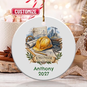 May include: A round white ceramic ornament featuring a yellow hard hat, blueprints, a compass, a gear, and books. The text reads ENGINEER, Build, Solve, Innovate, Anthony, and 2027. A gold string is attached to the top for hanging purposes.