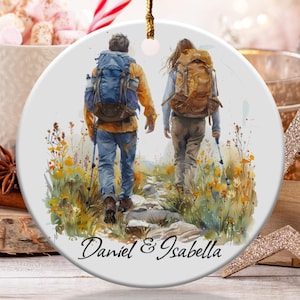 Puede incluir: Adorno redondo de cerámica con una ilustración de acuarela de dos excursionistas con mochilas caminando por un sendero. El adorno tiene una cuerda dorada para colgar y los nombres "Daniel & Isabella" están escritos en la parte inferior.