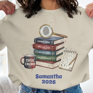 Könnte beinhalten: Beiges Sweatshirt mit einer Grafik von einem Stapel Bücher mit Titeln wie "Criminal Minds" und "Unsolved Cases", einer Lupe, einem Notizblock und einer Tasse mit der Aufschrift "True Crime". Der Name "Samantha" und das Jahr "2026" sind ebenfalls abgebildet.