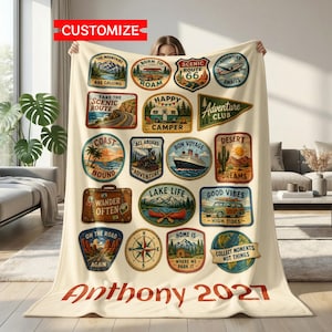 May include: A cream-colored fleece blanket featuring a grid of vintage-style travel patches with illustrations of mountains, campers, trains, ships, and landscapes. The text includes phrases like "Happy Camper," "Lake Life," and "Collect Moments Not Things," with the name "Anthony 2027" printed in large red letters at the bottom.