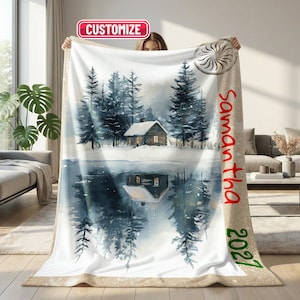 May include: A soft blanket featuring a watercolour-style winter scene with a cabin nestled among pine trees and its reflection in water. The design includes a tan side border with the red text Samantha and the green text 2027. A pink CUSTOMIZE label is visible.