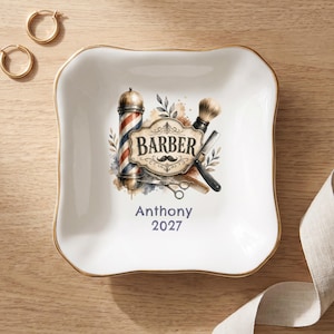 Peut inclure: Un vide-poche carré en céramique blanche avec un bord doré, orné d'un motif central représentant un poteau de barbier, un blaireau, un rasoir droit et un peigne. Le texte indique BARBER, Anthony et 2027, avec des touches d'aquarelle et des feuilles.