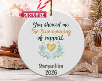 May include: A round white ceramic Christmas ornament with a gold hanging string. The ornament has the text "You showed me the true meaning of support." and a graphic of hands holding a gold heart. The name "Samantha" and the year "2026" are also printed.