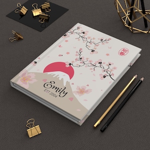 Op de afbeelding: Een wit notitieboek met een Japans geïnspireerd ontwerp met een rode zon, de berg Fuji en kersenbloesems. Het notitieboek is gepersonaliseerd met de naam "Emily" en het jaar "EST 2004". Gouden en zwarte potloden en gouden paperclips zijn ook zichtbaar.