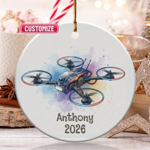 Peut inclure: Ornement de Noël rond en céramique blanche avec un dessin de drone. Le drone est illustré avec des détails bleus, orange et argentés. Le nom "Anthony" et l'année "2026" sont imprimés en dessous.